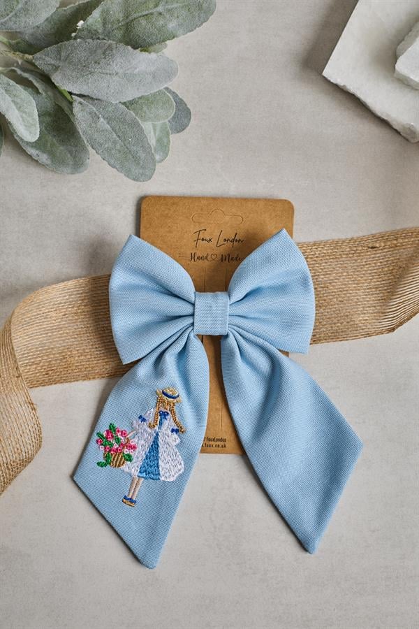 A blue embroidered bow tie with a floral pattern, displayed on a beige background, attached to a brown ribbon.