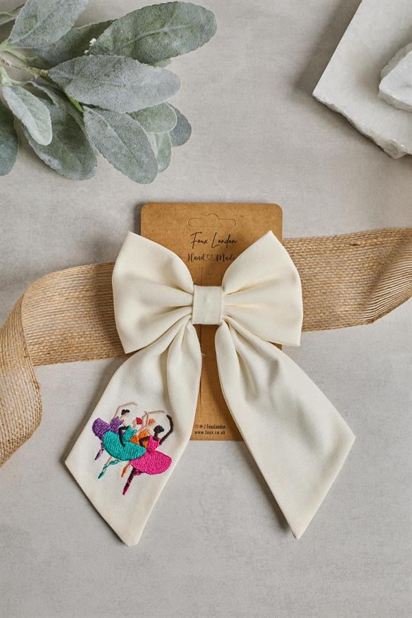 A beige bow tie with a ballerina embroidery on it, displayed on a cardboard with a plant and a small tag with the brand name.