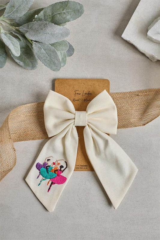 A beige bow tie with a ballerina embroidery on it, displayed on a cardboard with a plant and a small tag with the brand name.
