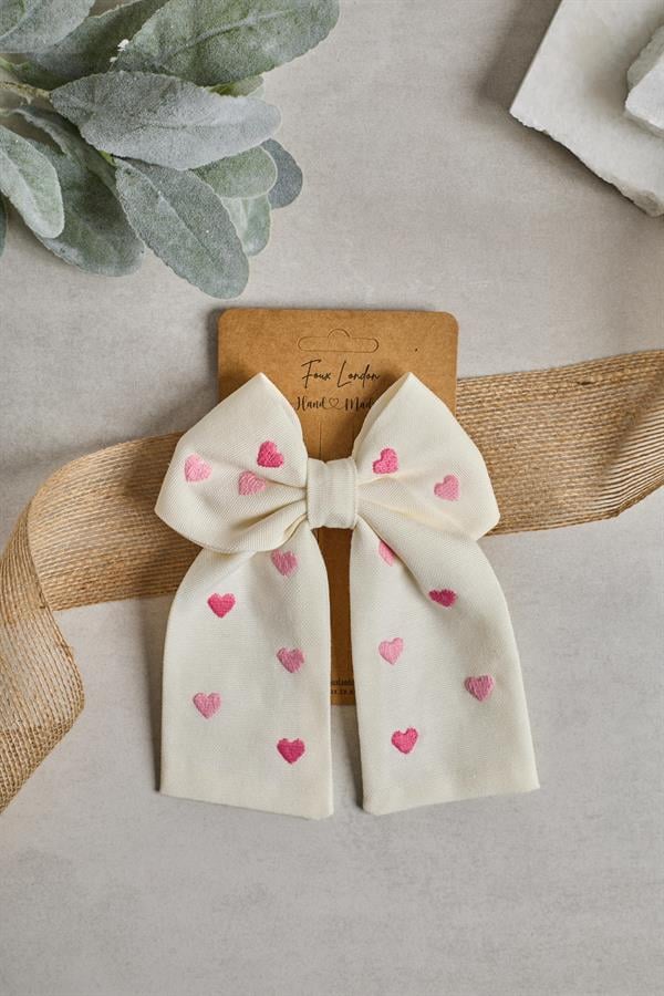 A beige bow tie with embroidered heart details, displayed on a card with a greeting message.